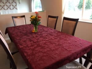 Large dining table with six chairs around it. It was a rust-colored table cloth covering it and a floral centerpiece.