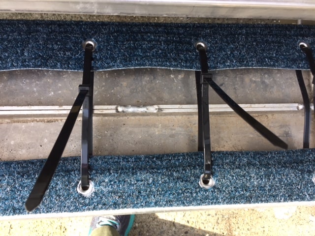 The underside of an RV step. The zip-tie used for attaching the carpet to the step is close up but the tails of the zip-ties are long and hanging off. 
