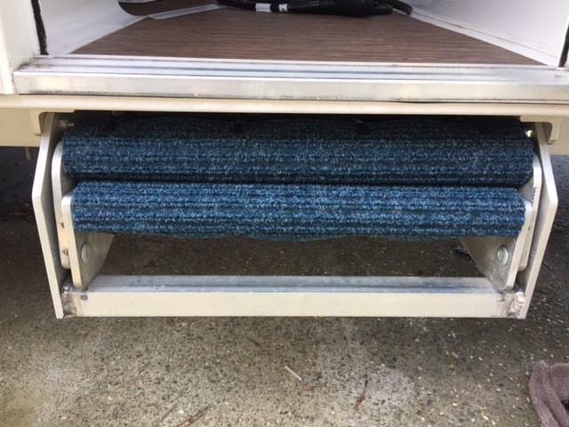 RV steps folded on themselves and tucked under the door of an RV. Mostly, the photo shows the metal of the steps and the thin line of blue carpet.