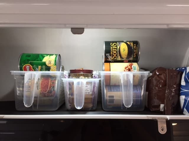 Three clear plastic pantry baskets with handles, filled with cans. They are sitting on a shelf in an RV.