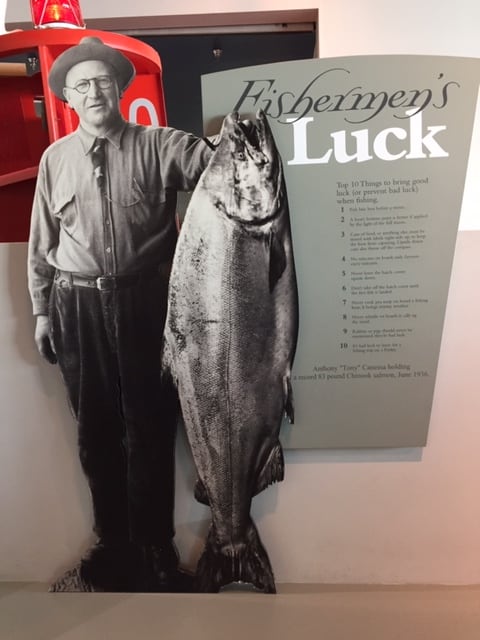 Photo of a man holding an 83 pound salmon he caught in 1936. The Fisherman's Luck list stands next to him.