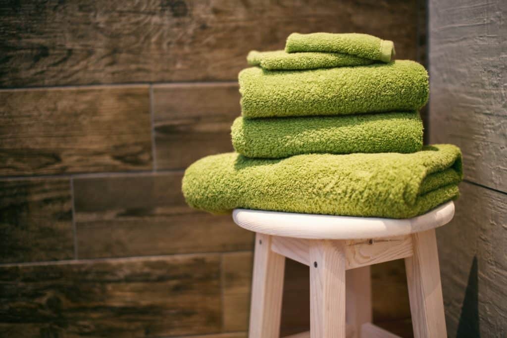 Photo by Denny Müller on Unsplash. Three towels and two wash clothes (all green) folded and sitting on a stool in the corner. 