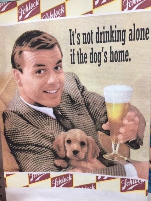 Sign with a man holding a beer and a puppy. It reads, "It's not drinking alone if the dog's home."