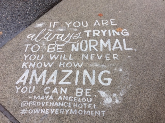 Painted in white on a sidewalk, "If you are always trying to be normal, you will never know how amazing you can be." Maya Angelou #OwnEveryMoment