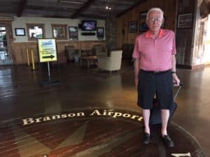 Man with white hair, red shirt and black shorts wheeling a piece of luggage. He is standing on the welcome sign at the Branson Missouri airport.
