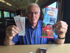 Older man with white hair holding two tickets and a brochure, sitting at a table.