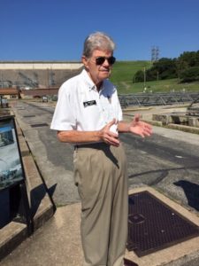 Older man with gray hair, sunglasses, a white shirt and tan pants, standing outside at a fish hatchery.