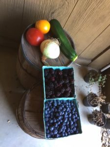 Sitting on wooden barrels: red tomato, yellow tomato, white onion, large zucchini, container of blueberries and a container of blackberries.