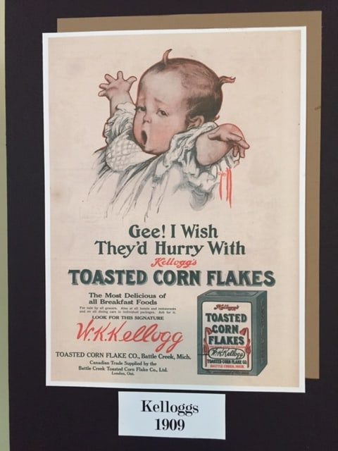 Advertisement from 1909. Shows a baby yawning at the top and a box of Kellogg's Corn Flakes in the lower corner. The caption reads, "Gee! I Wish They'd Hurry With Kellogg's Toasted Corn Flakes." 