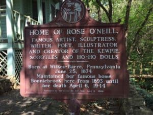Brown historical marker. It reads: Home of Rose O'Neill. Famous artist, sculptress, writer, poet, illustrator and creator of the Kewpie, Scootles and Ho-Ho dolls. Born at Wilkes-Barre, Pennsylvania June 25, 1974. Maintained her famous home Bonniebrook here from 1893 until her death April 6, 1944.