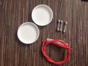 On a wood grain background, two white plastic lids, 3 small cylindrical fuses and a fuse hold with a red cord.