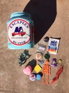 Light blue container with Escapees sticker and various trinkets that were inside the bucket including a orange plastic skeleton, a Wet One, a pink heart, pins and more.