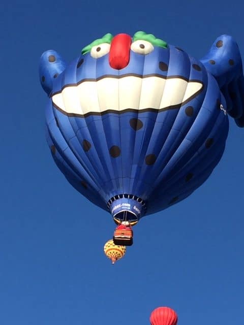 Dark blue hot air balloon with a big smiling face and ears. Two other hot air balloons are in the shot. All flying against a blue sky.
