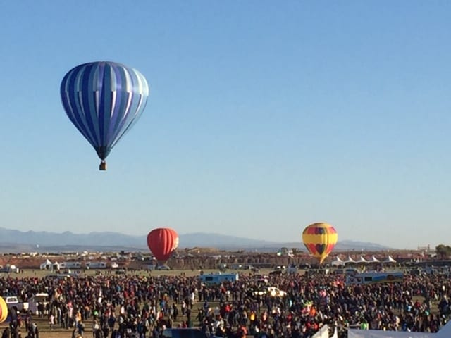 Blue and white hot air balloon in the air. Two hot air balloon inflated on the ground while lots of people wander around the launch field.