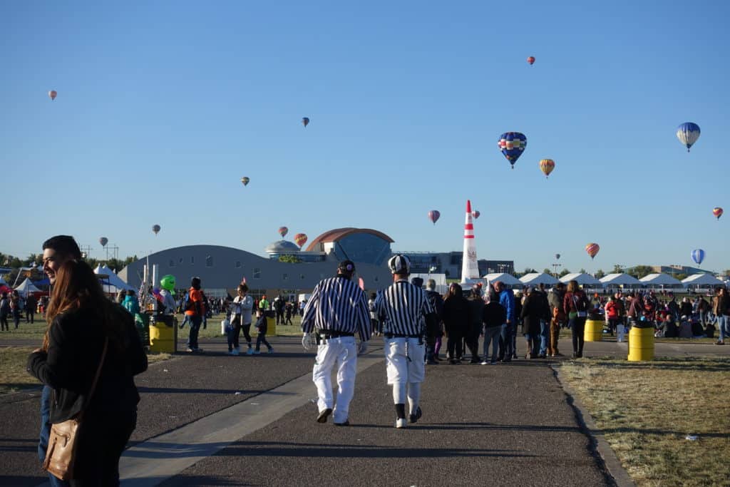 Hot air balloon against a blue sky with a crowd of people below, including two men in white pants with black and white striped shirts walking.