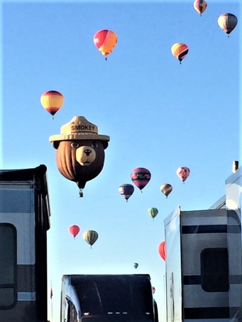 Smokey the Bear hot air balloon with lot of other balloons in the background flying over RVs.