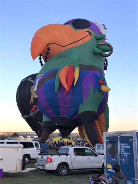 White trucks and trailers on the ground with a green parrot hot air balloon dressed like a pirate.