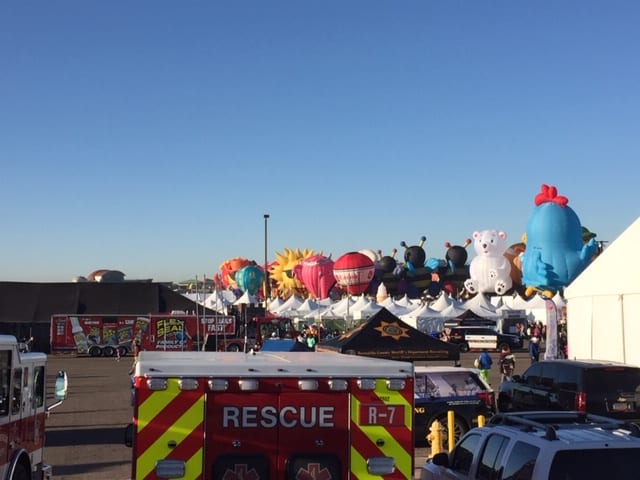 In the background are a line of inflated (but not launched) hot air balloons. In the foreground is people, ambulance and buildings