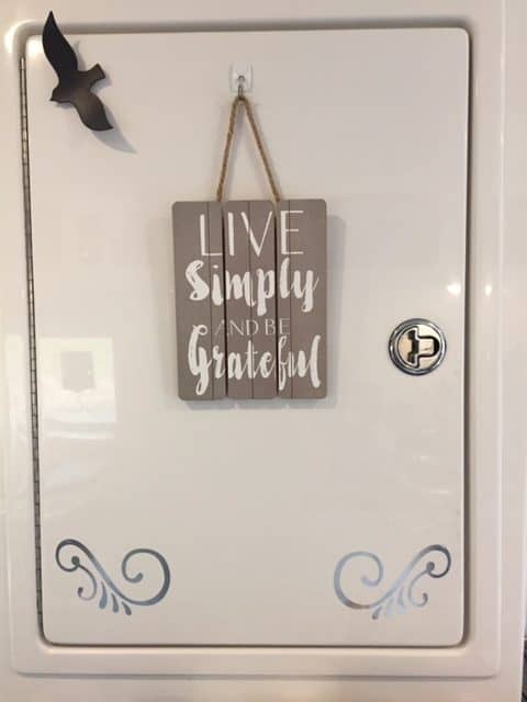 White whiny cabinet front with decorations--little black bird, silver swirls and a sign that read Live Simply and Be Grateful.
