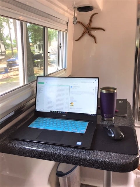 Taken inside an RV at the single-width dining table with windows looking out to trees. A computer, mouse, calendar and purple coffee mug are on the table.