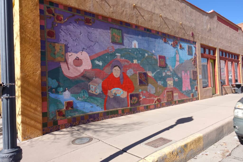 A mural painted on the side of a building with a woman cooking, a guitar and hills. 