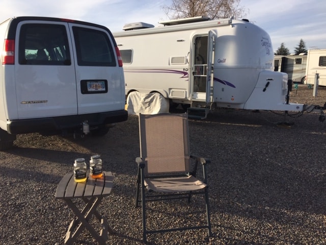 In the foreground is a tan camp chair and little table. There are two mason jars filled with moonshine on the table. In the background is a white travel trailer and a white van.