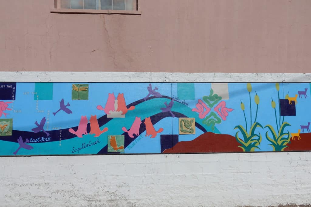A mural painted on the side of a building. light blue background with birds, wolves, deer and plants.