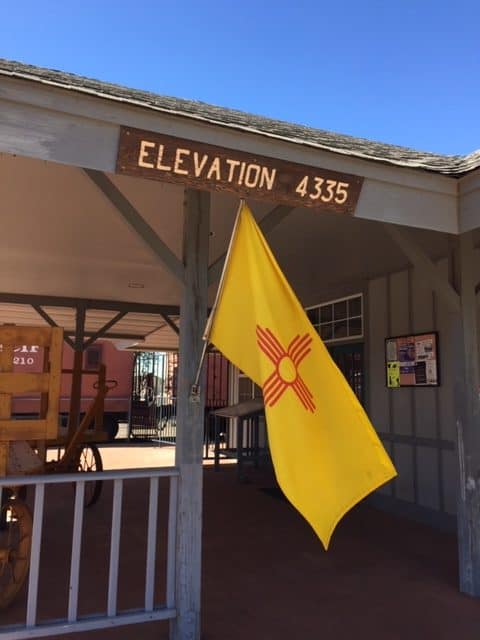 Front of the Visitors' Center in Deming, New Mexico. The state's yellow flag with red geometric design waves. There is also a sign that reads, "Elevation 4335."
