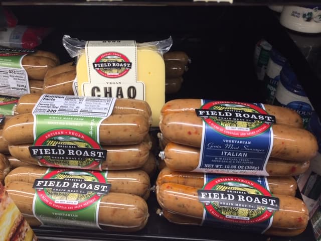 A grocery store deli case with a close up on packages of Field Roast vegan sausages and one package cheese slices tucked in.