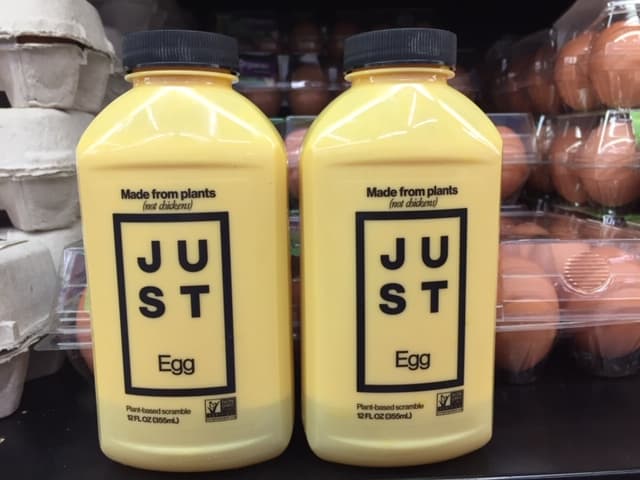 Two yellow containers of Just Egg. They are sitting on a grocery store shelf in front of cartons of eggs.