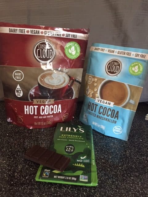 Two bags of Coconut Cloud hot chocolate--one is regular and the other is Toasted Marshmallow. Bags are standing up. Laying in front of the bags is a bag of Lily's Extremely Dark Chocolate with a piece of the chocolate sitting on top. All great vegan products.