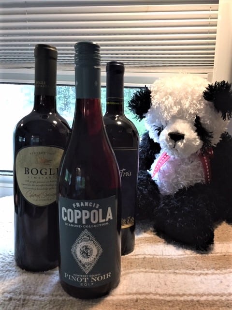 Three bottles of wine sitting on a blanket. A black and white stuffed teddy bear behind the bottles.
