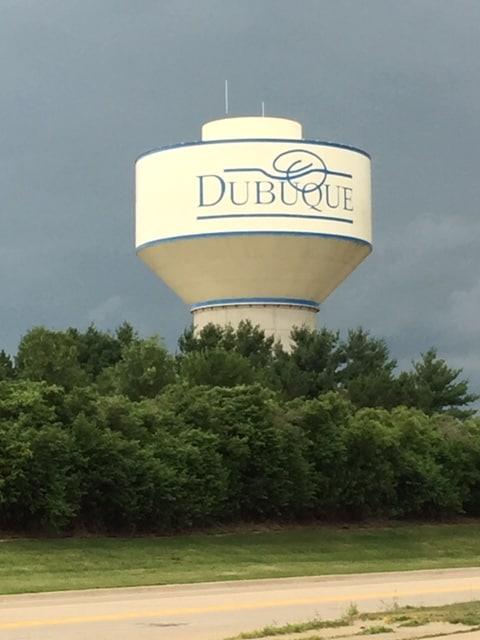 Water tower with the word Dubuque written in blue, coming up behind some trees and against a stormy sky.
