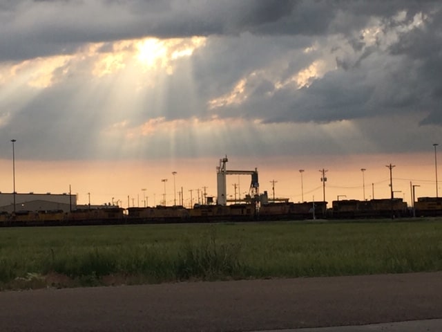 Silhouettes of trains and a lot of power poles. Dark clouds in the sky with the sun and sun rays coming through.