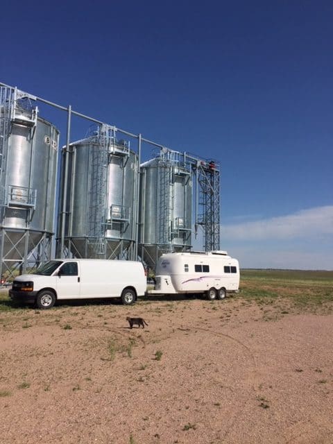 White van hooked up to a white trailer, parked on dirt in front of three tall silver grain silos with a dark blue sky in the background. And a cat in the foreground.