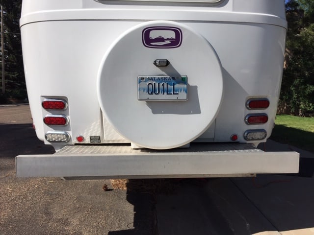 Back, lower half of a white travel trailer. Can see the bumper, the case for the spare tire as well as the blinkers.