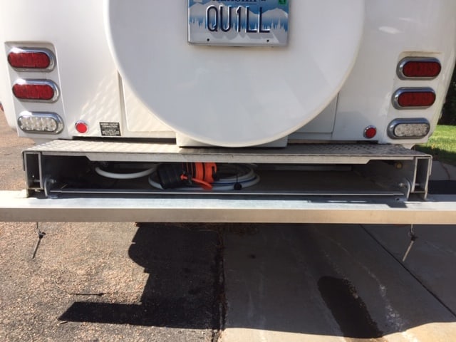 Back, lower half of a white travel trailer. Can see the bumper, the case for the spare tire as well as the blinkers. Bumper is opened and the white and blue garden hose with the sewer seal is tucked into it.