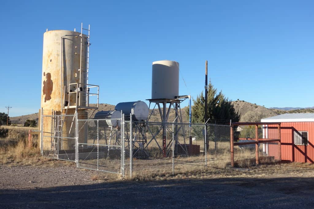 Behind chain-linked fence is 3 or four small water tanks and towers.