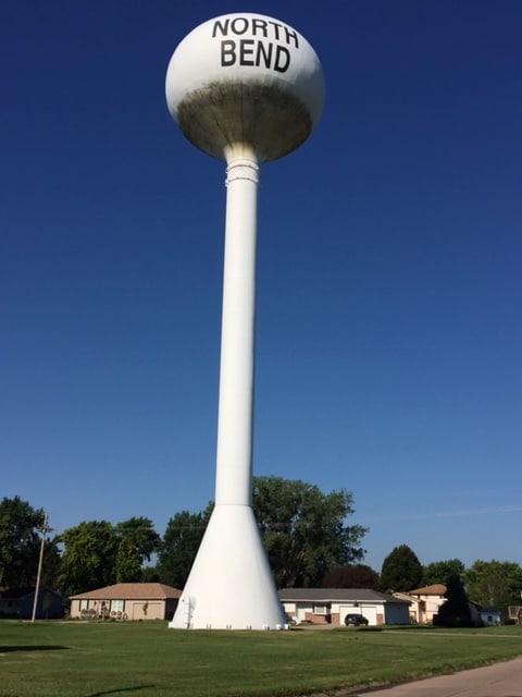 White water tower against a blue sky with the town's name, North Bend. Several houses with trees are behind it.