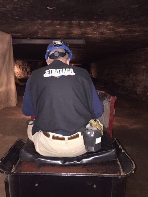 A man in a hard hat and a black shirt that reads, "Strataca." Photo is taken of his back as he sits on a cushion in the "conductors' position on a mine train.