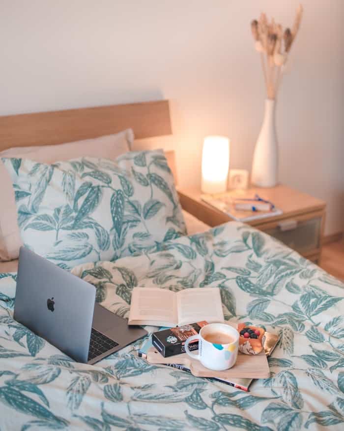 Computer, book,, pastry and cup of coffee on a bed with a green leaf design bedspread and matching pillow. Photo by Eea Ikeda on Unsplash
