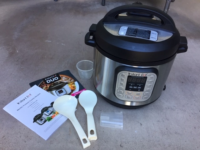 Instant Pot sitting on concrete. Next to it is two white plastic serving spoons, two plastic containers, an instruction manual and a recipe booklet.
