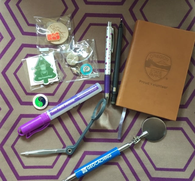 On a purple and gray geometrics clothes, geocaching supplies including a variety of small trinkets, pen, pencil, small leather bound notebook, tube of hand sanitizer, magnifying glass with tweezers attached, little round mirror on an extension pen.