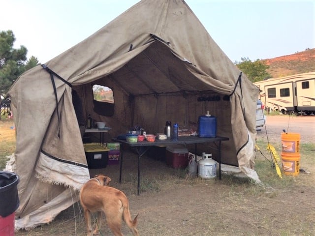 A large dirty white canvas tent set up at a campground. The doors are secured open opened. Inside is tables with food and cooking gear. A yellow dog is in the foreground of the photo.