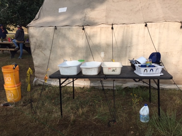 A table set up outside a large canvas tent with three white tubs and two dish drainers. A container of bleach sits under the table.