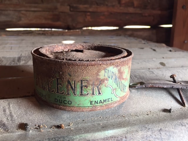 A small old almost entirely rusted can of furniture enamel. It sits in thick dust.