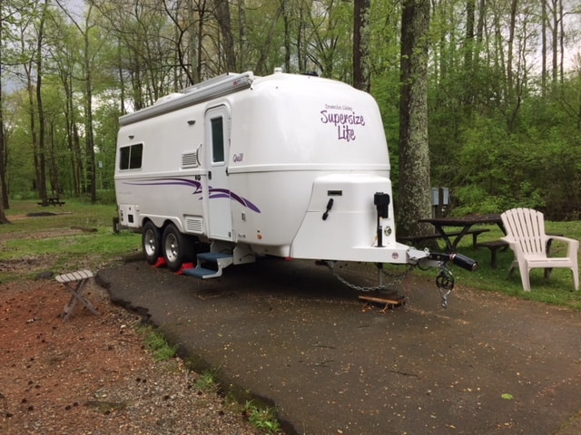 White Oliver Travel trailer parked on a strip of blacktop with lots of green trees all around.