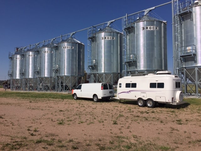 White van and trailer in front of six silver grain silos.