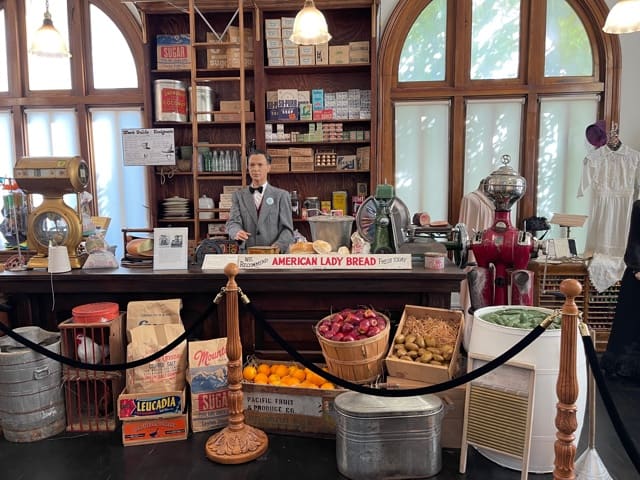 A full size display that looks like an old general store, including barrels of fruits and veggies and a man behind the counter.