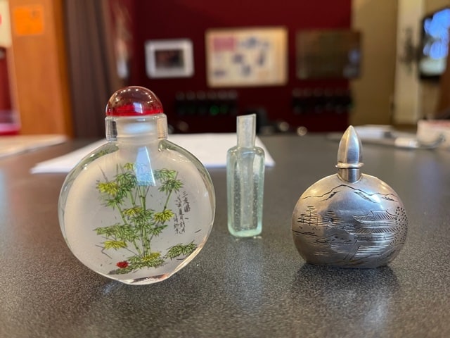 Three bottles. One is round and flat with a decorative tree on it. On is a tiny square on. Last one is a metal round one.